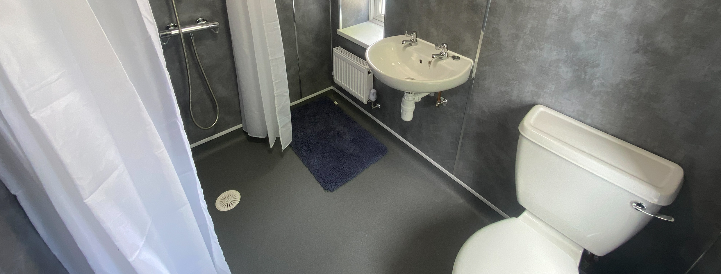 The view is looking from high up in the wet room and looking down toward the floor. The walls are grey with a slightly darker grey flooring. There is a shower curtain prominent in the image to the left, with a toilet on the right hand side and a sink toward the middle of the image on the wall.